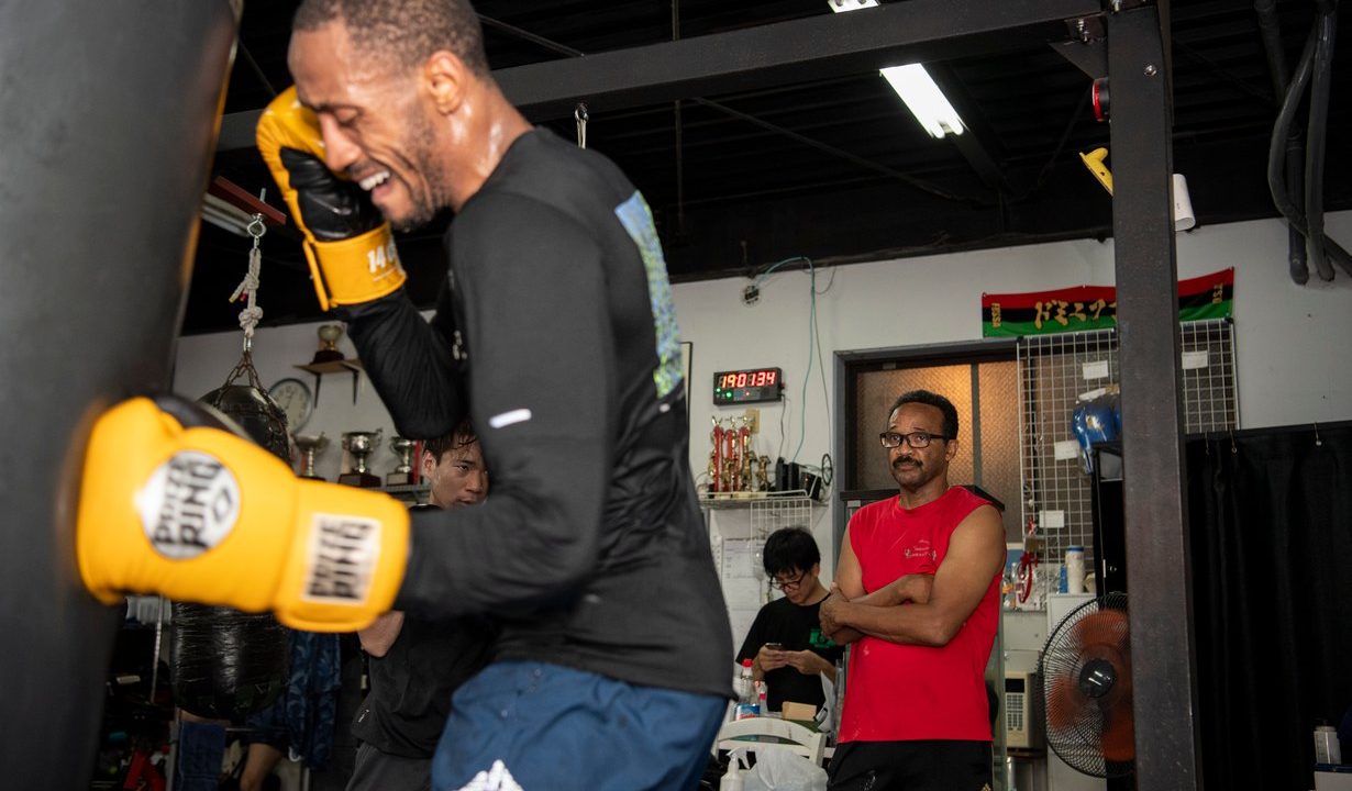 Boxer schlägt in einem Boxgym auf einen Sandsack, im Hintergrund trainiert ein weiterer Sportler