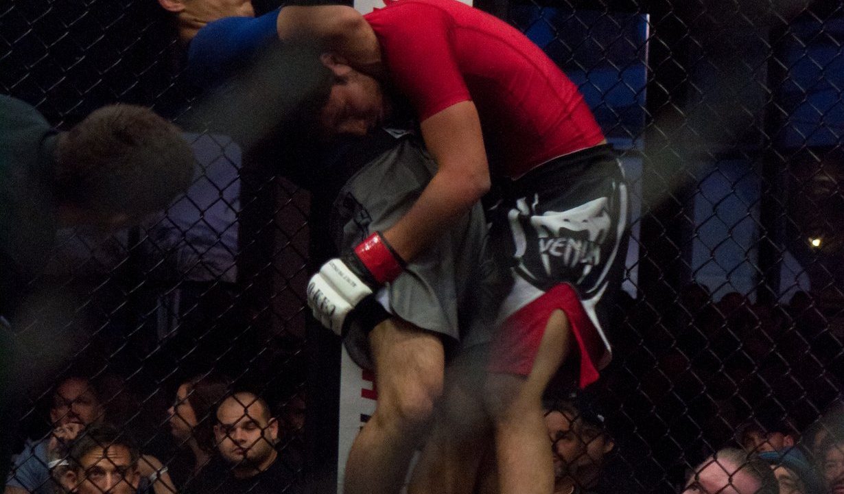 Zwei MMA-Kämpfer im Käfig, einer setzt einen Würgegriff an, Zuschauer hinter dem Gitter
